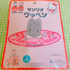 May include: A vintage Sanrio Marron Cream patch on a pink background. The patch features a gray bunny character with a white collar, surrounded by pink flowers and the text "Marron Cream". The packaging includes Japanese text and illustrations of Hello Kitty and other characters.
