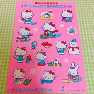 May include: A sheet of Hello Kitty stickers on a pink background. The stickers feature Hello Kitty in winter scenes, skiing, and with a snowman. Text includes "HELLO KITTY" and Japanese text. The sheet also has snowflakes and a bunny.