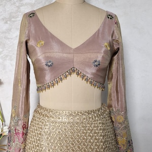 Puede incluir: Una blusa malva de manga larga con escote en V profundo y bordados florales intrincados. La blusa presenta delicadas cuentas en el escote y se combina con una falda dorada bordada. Las mangas tienen patrones coloridos detallados.