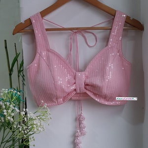 May include: A pink sequined crop top with a bow detail and a tie closure. The top has a deep V-neckline and is decorated with small, white sequins. The top is on a hanger and has a white background.