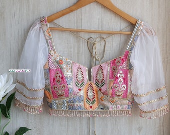 May include: A white crop top with sheer puff sleeves and a colorful patchwork design. The top has a deep V-neckline and a tie closure at the back. The fabric is embellished with sequins and beads.