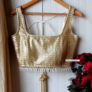 Könnte beinhalten: Ein goldfarbenes, mit Pailletten besetztes Crop-Top mit weißem Besatz und Perlenfransen. Das Top hat einen quadratischen Ausschnitt und einen Rückenschluss mit Schleife.