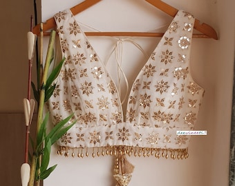 Puede incluir: Una blusa sin mangas de color blanco roto con un escote en V profundo, adornada con bordados florales dorados y detalles de espejos reflectantes. El dobladillo tiene flecos de cuentas doradas y una borla central. El texto deevineeti es visible.