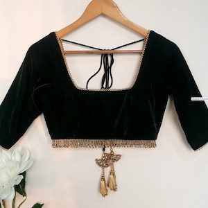 May include: A dark green velvet blouse featuring a square neckline, three-quarter length sleeves, and gold beaded trim along the edges. The garment includes a tie-back closure with dangling gold tassels and decorative accents at the waist.