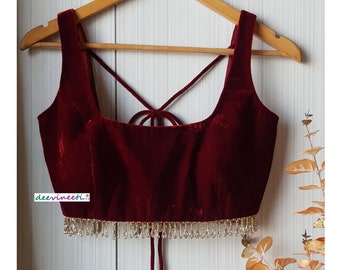 May include: A sleeveless deep red velvet crop top featuring a square neckline, thin crisscross back straps, and a dangling clear crystal bead fringe along the bottom hem. The garment is displayed on a wooden hanger with the text deevineeti visible.