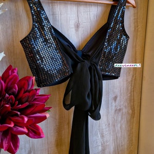 May include: A black sequined crop top with a black chiffon bow tie in the back. The top is hanging on a wooden hanger.