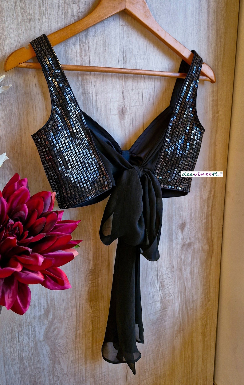 May include: A black sequined crop top with a black chiffon bow tie in the back. The top is hanging on a wooden hanger.