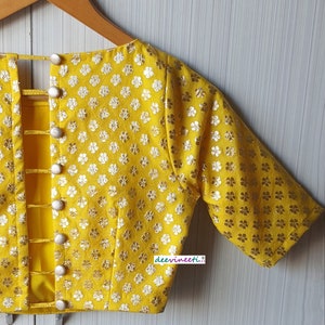 Pu&ograve; includere: Una blusa gialla con motivo floreale e chiusura con bottoni sul retro. La blusa ha maniche corte e scollo rotondo.