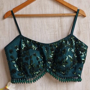 May include: A dark teal camisole top with thin straps, embellished with sequins and beads. The top features a floral design and a beaded fringe along the bottom edge. It is hanging on a wooden hanger against a light wood background.