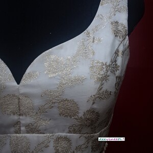 May include: A white sleeveless blouse with a gold floral pattern and a beaded trim. The blouse has a deep V-neckline and is made of a silky fabric. The text "deevinneeti..." is visible on the bottom right corner of the image.