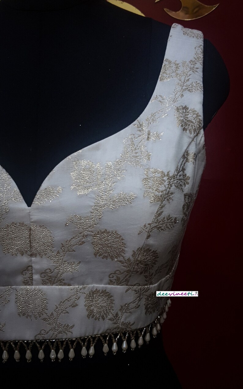 May include: A white sleeveless blouse with a gold floral pattern and a beaded trim. The blouse has a deep V-neckline and is made of a silky fabric. The text "deevinneeti..." is visible on the bottom right corner of the image.