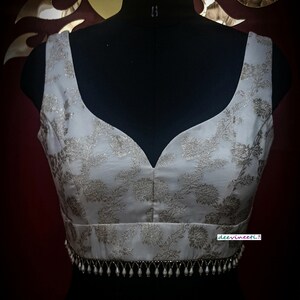 May include: A white sleeveless blouse with a silver floral pattern and a beaded trim around the bottom. The blouse has a deep V-neckline.