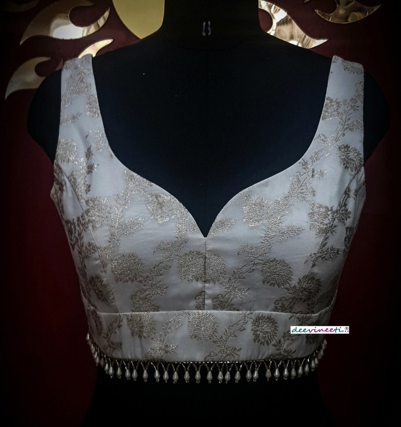 May include: A white sleeveless blouse with a silver floral pattern and a beaded trim around the bottom. The blouse has a deep V-neckline.