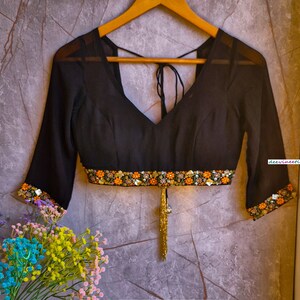 May include: A black sheer blouse with a V-neckline and a gold tassel. The blouse has a band of colorful embroidery around the waist and the sleeves.