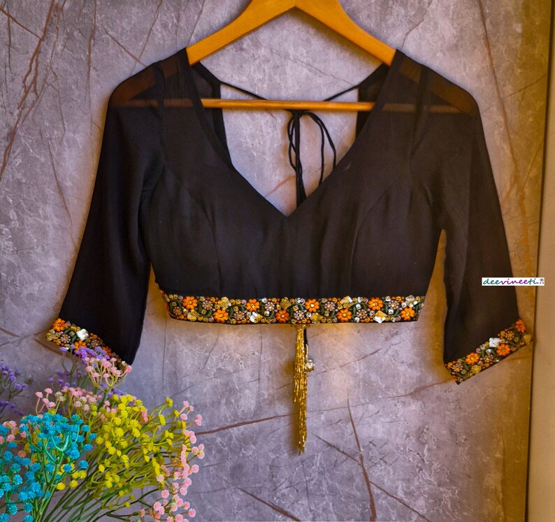 May include: A black sheer blouse with a V-neckline and a gold tassel. The blouse has a band of colorful embroidery around the waist and the sleeves.