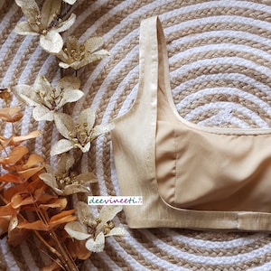 May include: A gold-colored, sleeveless, satin blouse with a scoop neck and a back zipper closure. The blouse is lying on a woven straw mat with a white and brown pattern.