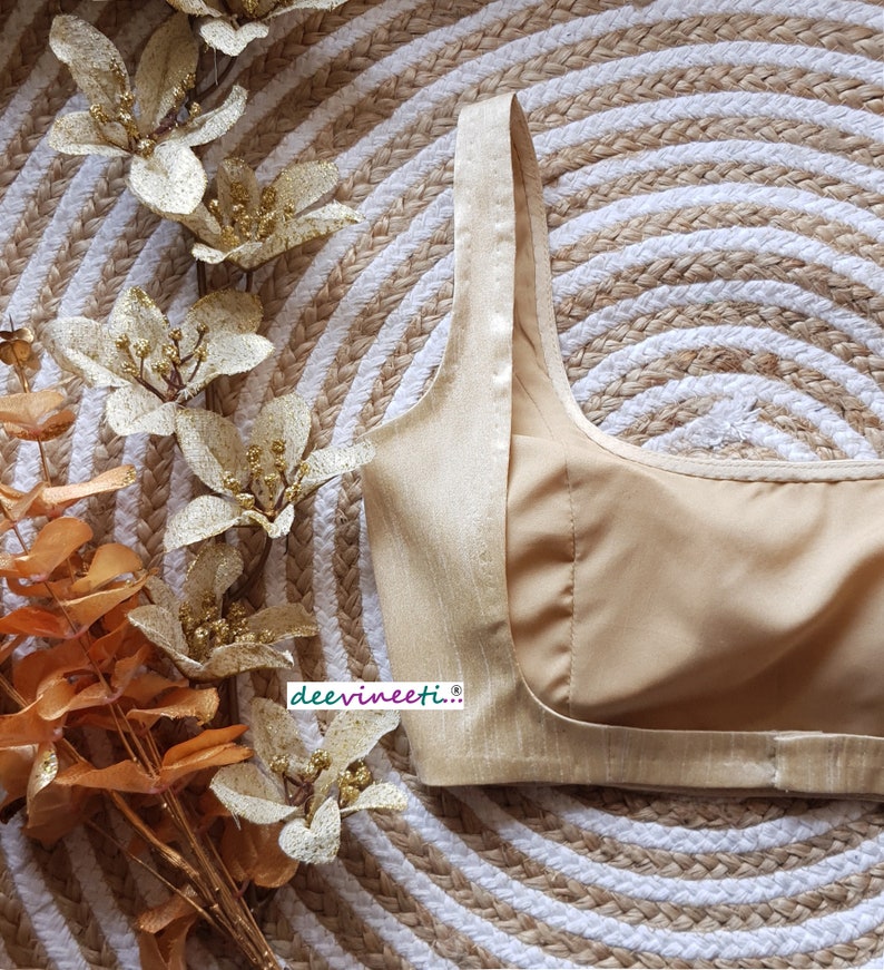 May include: A gold-colored, sleeveless, satin blouse with a scoop neck and a back zipper closure. The blouse is lying on a woven straw mat with a white and brown pattern.