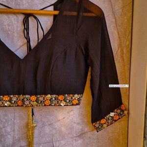 May include: A black sheer blouse with a V-neckline and embroidered trim. The trim features floral designs in orange, pink, and green. The blouse has a tie closure at the back and a gold tassel.