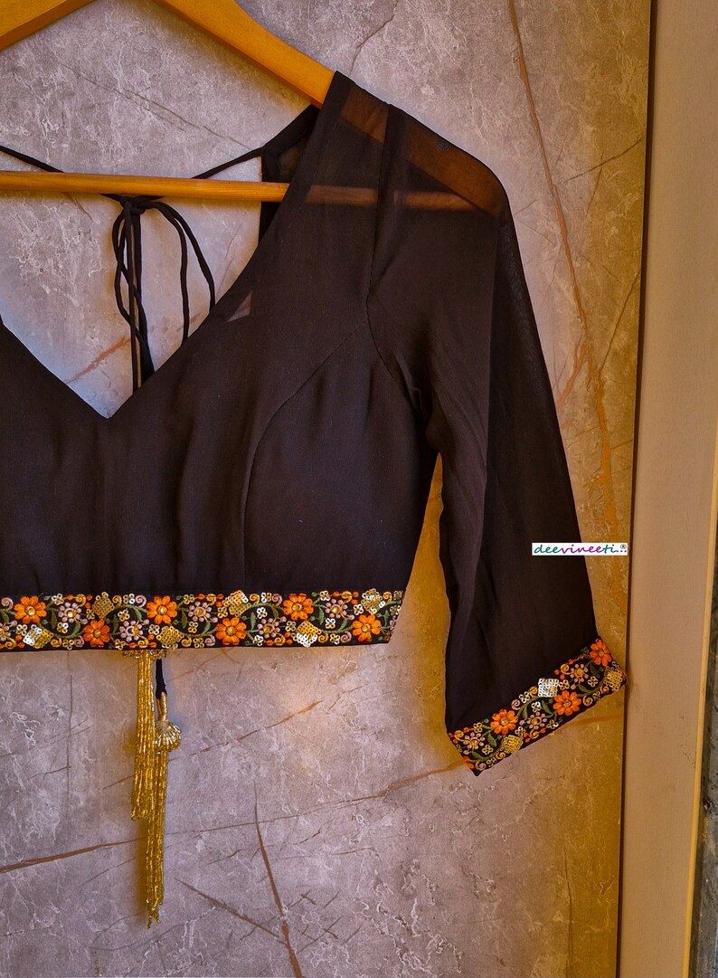 May include: A black sheer blouse with a V-neckline and embroidered trim. The trim features floral designs in orange, pink, and green. The blouse has a tie closure at the back and a gold tassel.