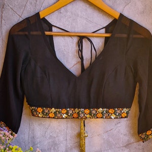 May include: Black sheer blouse with a V-neckline and embroidered trim. The blouse has a tie at the back and a tassel.