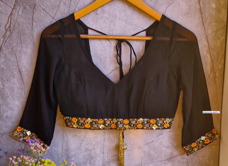 May include: Black sheer blouse with a V-neckline and embroidered trim. The blouse has a tie at the back and a tassel.