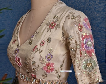 May include: A cream-coloured embroidered blouse with a V-neckline and short sleeves. The blouse is decorated with floral embroidery and sequins in shades of pink, purple, and green. The blouse has a beaded trim around the neckline and sleeves.