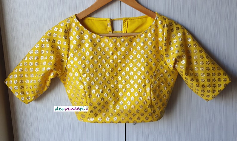Pu&ograve; includere: Blusa gialla con motivo floreale e maniche corte. La blusa ha una scollatura rotonda e un'apertura a chiave sul retro.