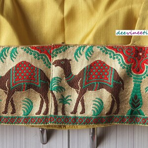 May include: A yellow fabric with a gold trim featuring a repeating pattern of camels and palm trees. The trim is decorated with red, green, and gold accents.