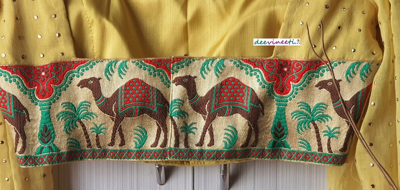 May include: A yellow fabric with a gold trim featuring a repeating pattern of camels and palm trees. The trim is decorated with red, green, and gold accents.