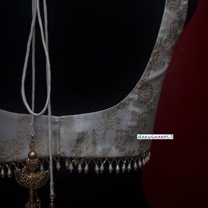 May include: A white embroidered blouse with a gold floral pattern and a white beaded trim. The blouse has a white string tie at the back with gold tassels.