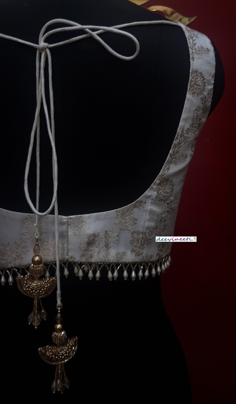 May include: A white embroidered blouse with a gold floral pattern and a white beaded trim. The blouse has a white string tie at the back with gold tassels.