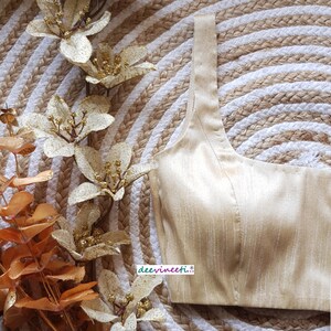 May include: A gold-colored, sleeveless, satin blouse with a square neckline. The blouse is on a woven, circular, beige and white background.