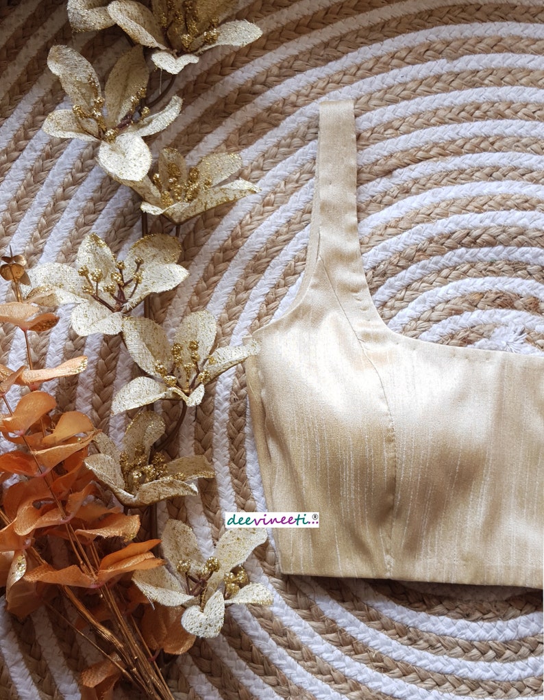 May include: A gold-colored, sleeveless, satin blouse with a square neckline. The blouse is on a woven, circular, beige and white background.
