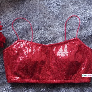 Puede incluir: Un top corto rojo de lentejuelas con tirantes finos. El top está hecho de una tela brillante con un brillo sutil. Las correas son ajustables y el top tiene un escote redondo.