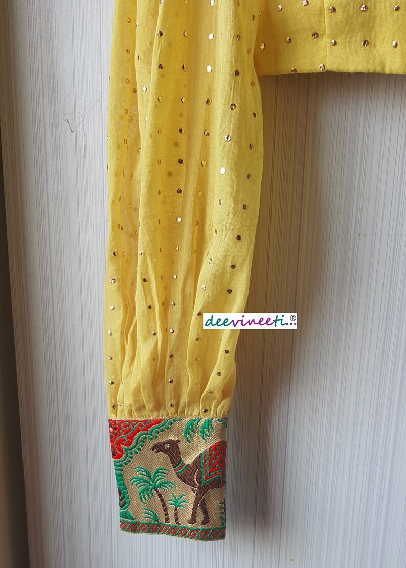 May include: Yellow sheer blouse with gold sequins and a colorful embroidered cuff featuring a camel and palm trees. The cuff has red, green, and gold accents. The text "deevineeti..." is printed on the blouse.