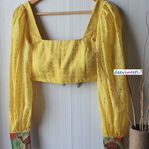 May include: A yellow crop top with a square neckline and long sleeves. The top is decorated with small gold sequins and has a printed design on the cuffs. The text "dee vineeti..." is visible on the right side of the image.