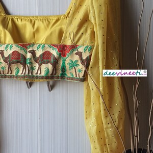 May include: A yellow crop top with a camel print trim and puff sleeves. The sleeves have gold sequins. The top is on a hanger against a white wall.