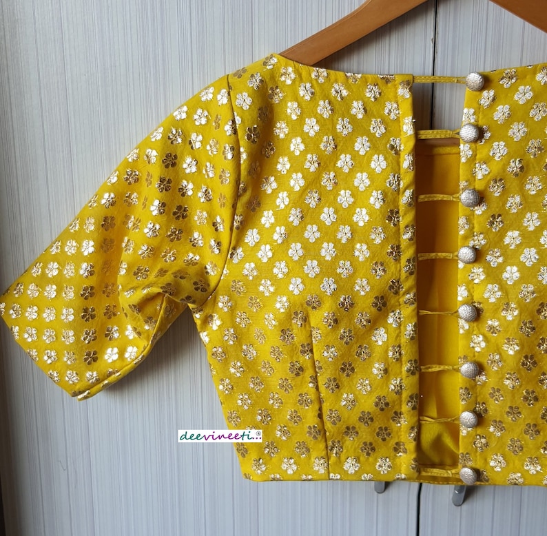 Pu&ograve; includere: Una blusa gialla con motivo floreale e un design posteriore con una serie di chiusure a bottone.