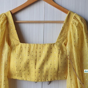 May include: A yellow crop top with a square neckline and puff sleeves. The top is decorated with small gold sequins.