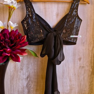 May include: A black sequined crop top with a black chiffon bow tie in the back. The top is hanging on a wooden hanger against a woodgrain background.