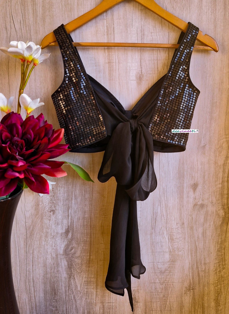 May include: A black sequined crop top with a black chiffon bow tie in the back. The top is hanging on a wooden hanger against a woodgrain background.