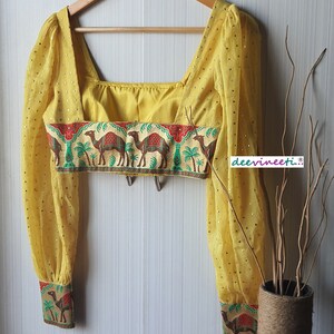 May include: A yellow crop top with long sleeves and a square neckline. The top has a band of fabric with a camel print around the waist and cuffs. The fabric is sheer and has small gold dots.
