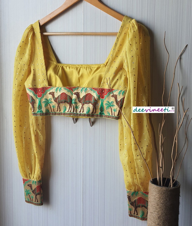 May include: A yellow crop top with long sleeves and a square neckline. The top has a band of fabric with a camel print around the waist and cuffs. The fabric is sheer and has small gold dots.