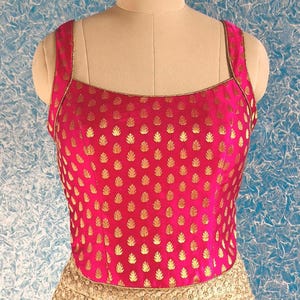 May include: A hot pink, sleeveless top with a square neckline and gold leaf pattern. The top has gold trim along the neckline and straps. The bottom portion of the garment is a light gold color with a floral design.