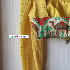 May include: Yellow crop top with a patterned trim featuring camels and palm trees. The top has a scoop neck and long sleeves with a gathered puff at the shoulder. The sleeves are embellished with gold sequins.