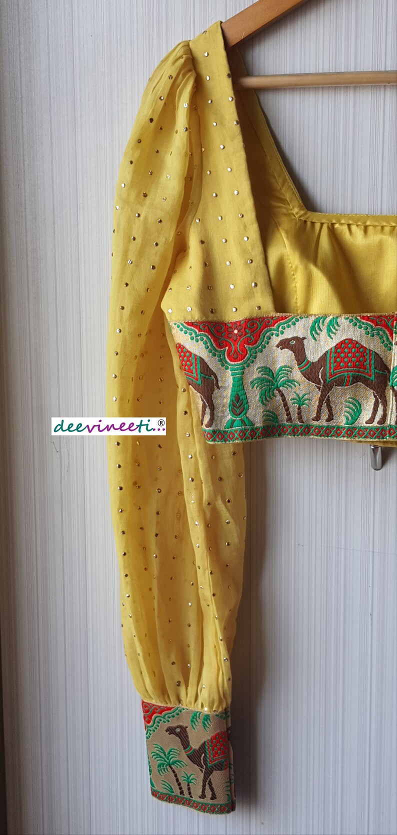 May include: Yellow crop top with a patterned trim featuring camels and palm trees. The top has a scoop neck and long sleeves with a gathered puff at the shoulder. The sleeves are embellished with gold sequins.