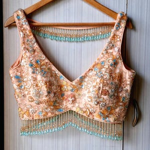 May include: A sleeveless peach-colored blouse featuring a deep V-neckline, heavily embellished with intricate gold, silver, and light blue sequins and beads. The garment includes a dangling fringe of light blue teardrop beads at the hem.