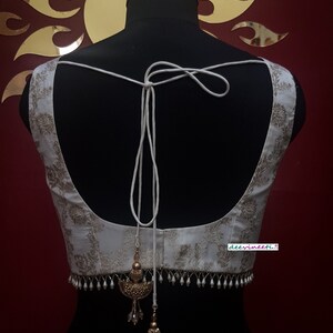 May include: White sleeveless blouse with a gold floral pattern and a white tassel tie at the back. The blouse has a beaded trim with gold accents.
