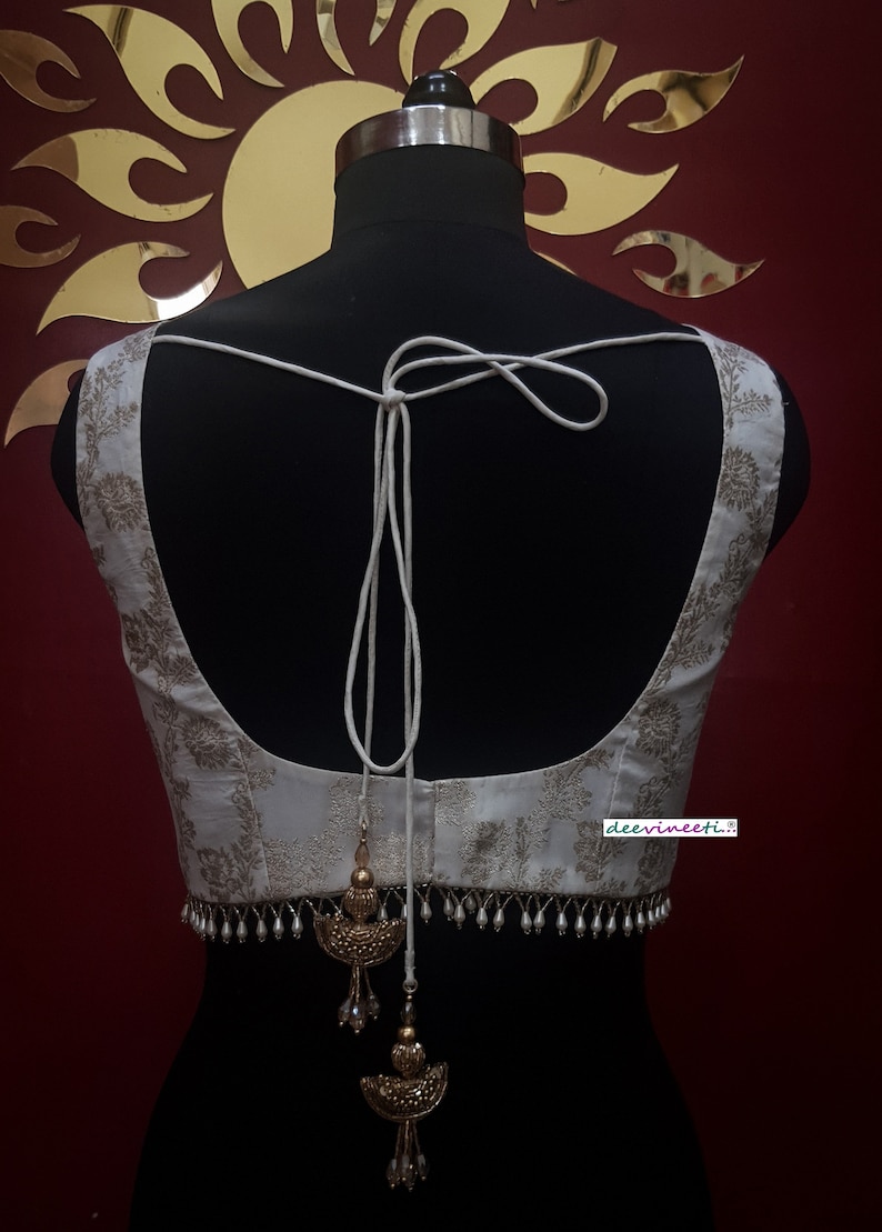 May include: White sleeveless blouse with a gold floral pattern and a white tassel tie at the back. The blouse has a beaded trim with gold accents.