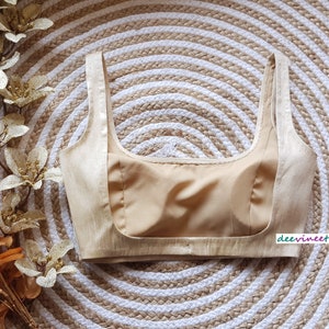 May include: A beige sleeveless blouse with a satin-like finish. The blouse has a scoop neckline and a fitted bodice.